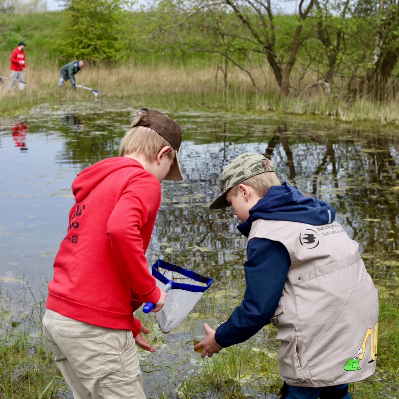 Bilden visar två barn som står vid kanten av en vattensamling, möjligen en sjö eller damm. Det ...