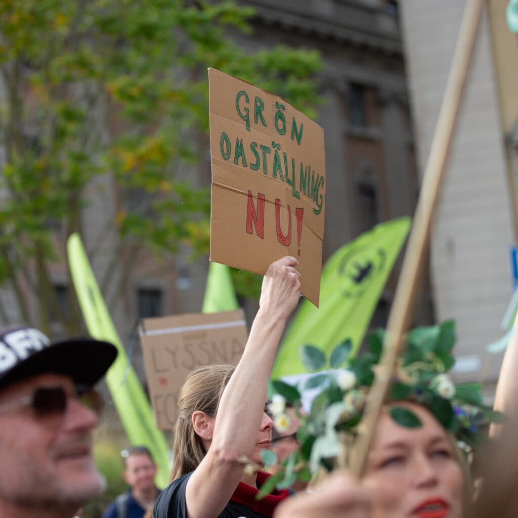 Bilden visar en demonstration där människor håller upp skyltar. En av skyltarna har texten "GRÖN ...