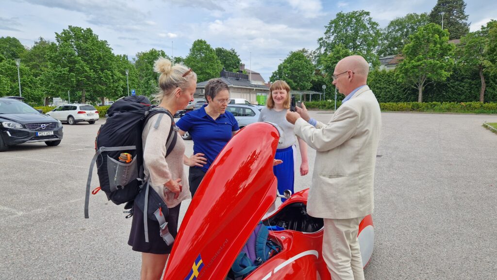 4 personer står runt en röd velomobil. En man i kostym intervjuar en kvinna i blå piké.